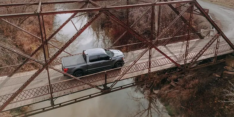 Aerial view of gray pickup crossing narrow rusted truss bridge over river, showcasing truck tires on rural road