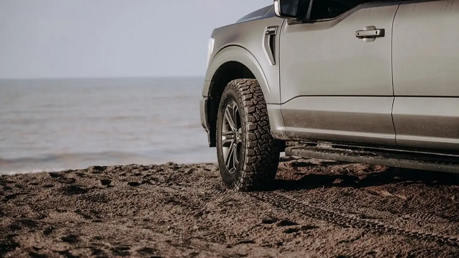 SUV with all terrain tires parked on sandy beach near calm waters, ready for adventure